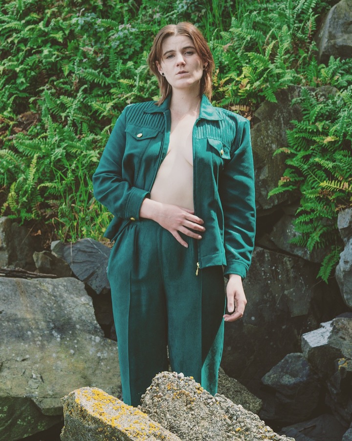 Me in some rocks wearing a green jumpsuit and bright green furns in the background.