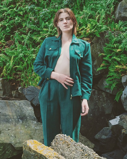 Me in some rocks wearing a green jumpsuit and bright green furns in the background.