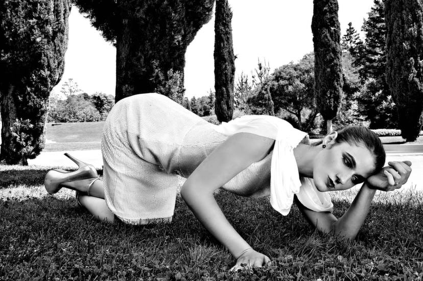 Black and white, grass, tall hedge..trees? In background. White dress, texture, draping. Hair pulled back with shiny shoes. Kneeling, err more on the ground. Table position in yoga? But in deep thought. Hmmm.