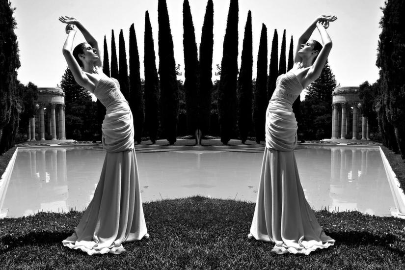 Black and white mirrored image, standing on grass with arms up leaning back in front of a super big fountain pool with more hedge trees and big columns, sleeveless dress going down onto the grass. Draaaap.