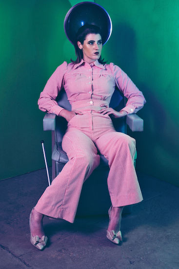 Photo of me in hairdrying chair with green background in red pinstripe wide leg trousers and button up crop top with collar and long sleeves, gold heels, looking left