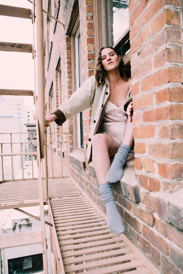 Sitting in window holding on to rails of a fire escape in the City. Brick. Grey socks, beige coat with brown corduroy cuffs. Light pink silk one piece.